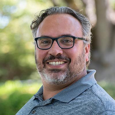 Portrait of Alex Paez, smiling, wearing glasses and a gray polo